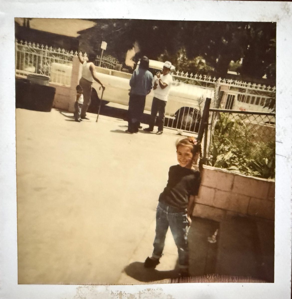 Early 90s in Compton, CA.  Little Karina is smiling big for the camera and is seen wearing a black tee shirt with cuffed blue jeans while standing outside of her childhood home. To her right is her garden. In the background are her now-deceased father, two uncles, and her small cousin.