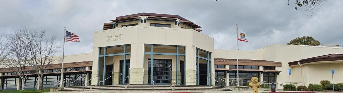 Octogon-shaped building entrance for BUTTE COUNTY COURTHOUSE and the building is pale yellow with blue beams in front of a dark and stormy-looking sky. The entrance is surrounded by stairs and flanked by two flag poles, with an American flag raised on the left and the California state bear flag raised on the right.