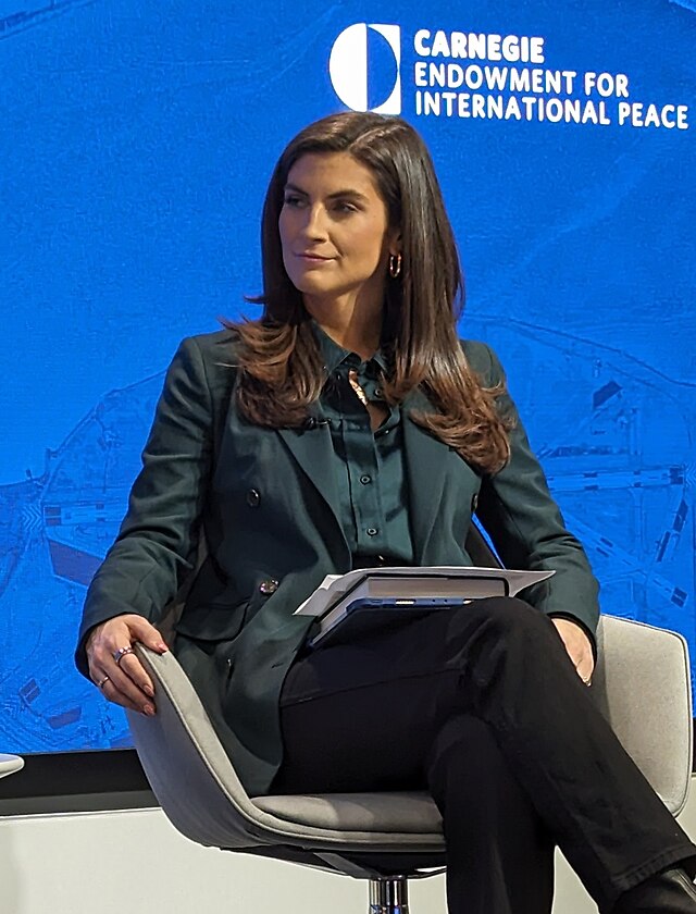 Kaitlin Collins sitting stoic in a chair, wearing a navy blue blazer and long black pants.