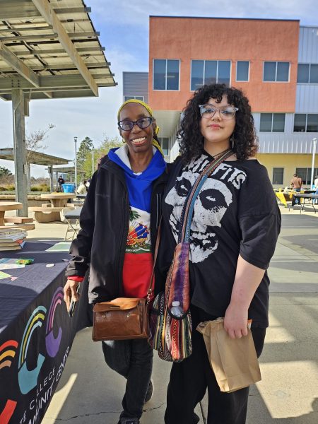 Two women stand closely posing in front of a booth table and tall, colorful office building and are being shaded by solar patio covering from direct sunlight.
