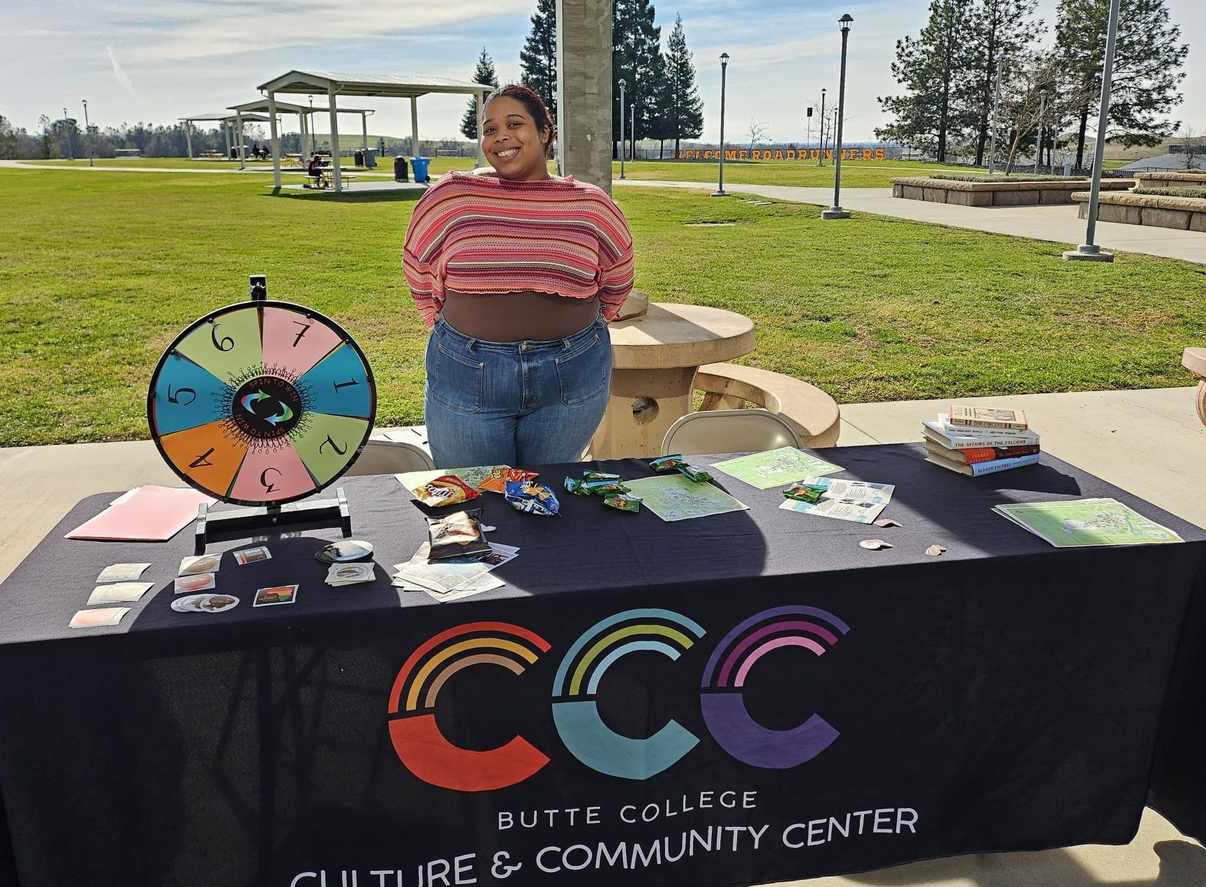 Young woman smiles behind a booth labeled “Butte College Culture & Community Center” decorated with flyers and a game wheel as sun shines on a large quad with grass behind her.