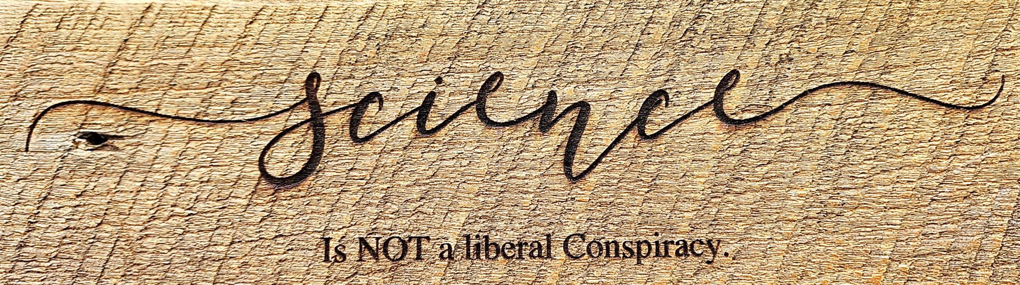 A woodburned engraving on roughly planed oak wood reads "Science - Is NOT a Liberal Conspiracy". In decorative cursive "Science" is engraved with swirls reaching outward from the first and last letters, "S" and "E". Much smaller underneath follows, "Is NOT (all capitalized) a liberal Conspiracy", with a capital "C" for consipracy.