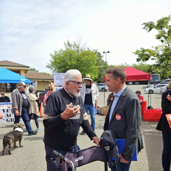 A man with white hair leans on a walker chair as he speaks to a man in jeans and a sports jacket. The man with the walker holds his right hand up as he speaks with the younger man in the sports jacket. The men stand in front of pop-up tents and others wearing large hats and sunglasses as the sun casts a long shadow in the afternoon light.