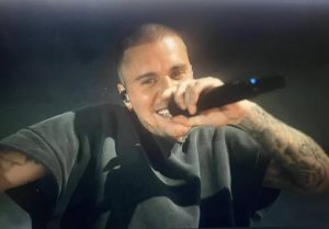 Justin Bieber with mic in hand, performing on stage at Coachella, with a black T shirt, and rocking a shaved head.