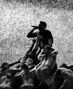 A black and white photo of Trent Reznor with dancers, climbing on him, while he sings