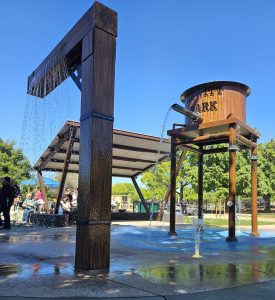 Fancy new construction of a water tower with words “HEWITT PARK” and a dripping train post stand tall over fountains spraying up water pooling in reflections on the concrete as families sit close by in the shade.