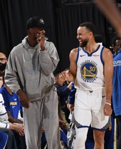 Steph Curry in Golden State Warriors uniform on the right, standing next to Jimmy Butler in a grey sweatsuit, to the left. 