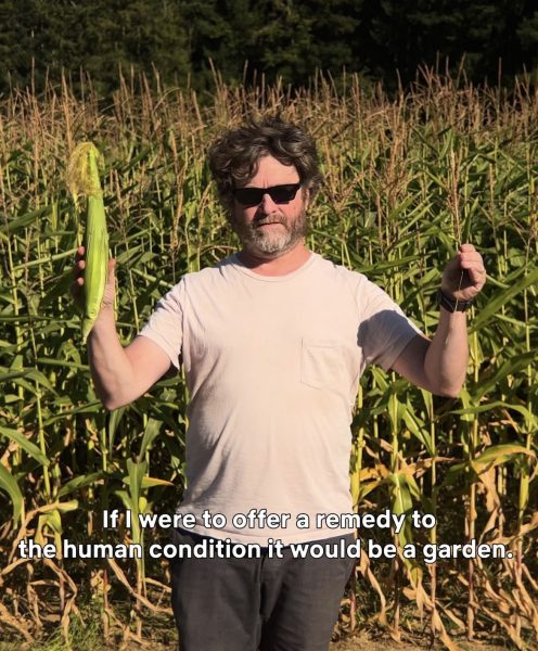 Host holding a piece of corn, in front of a corn filed. wearing a white shirt and black sun glasses.