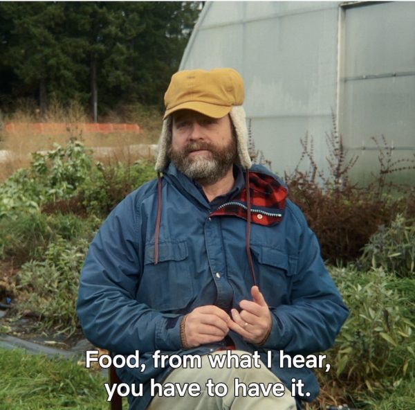 A screenshot of the host talking about food, sitting in a chair, with plants and a green house in the background. He's wearing a yellow hat and a blue jackey with some red flannel.